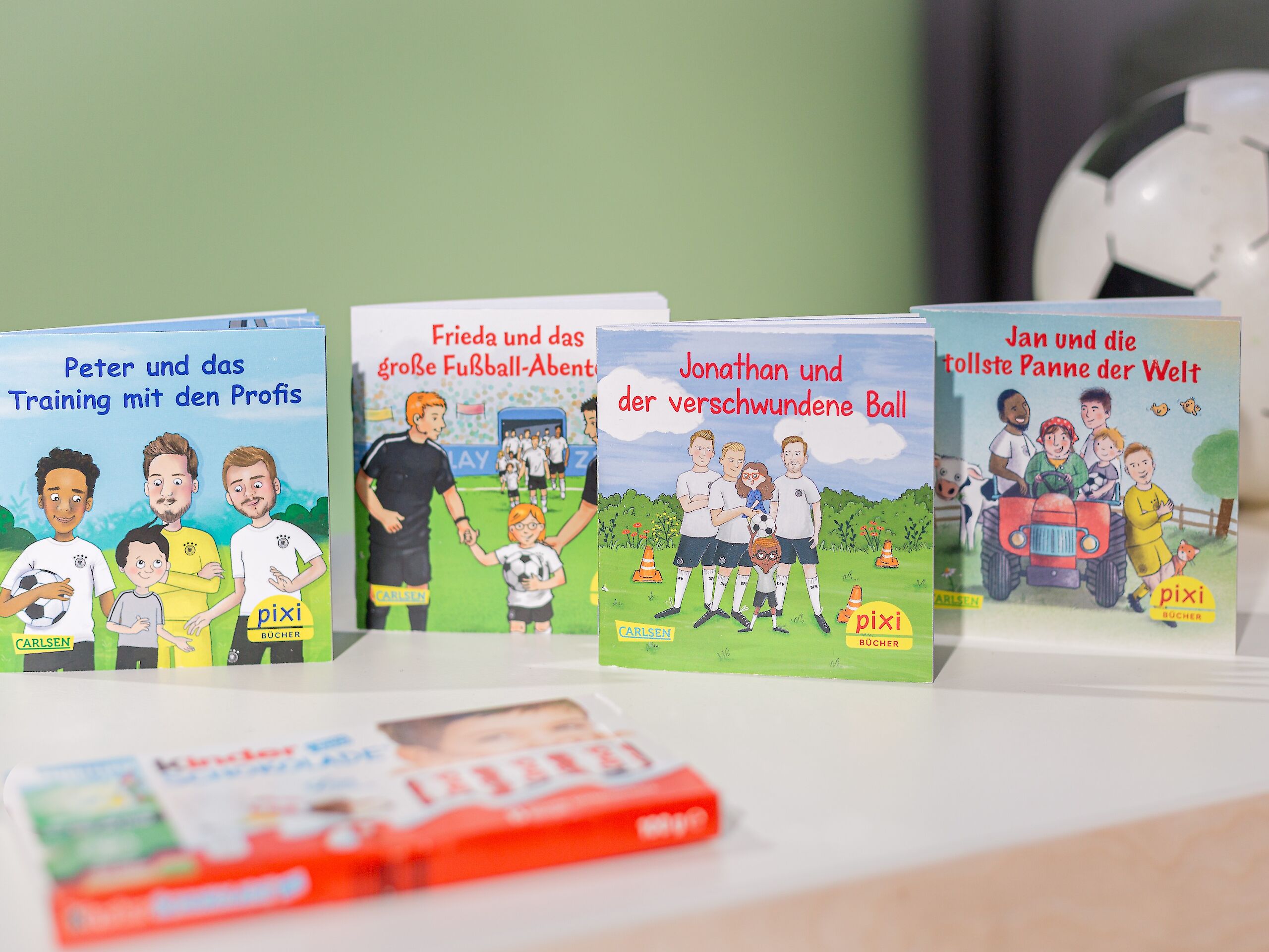 Das Bild zeigt vier Pixi-Bücher mit Fußballgeschichten, die auf einem Tisch stehen. Im Hintergrund liegt ein Fußball. Im Vordergrund liegt eine Packung Kinder Schokolade.