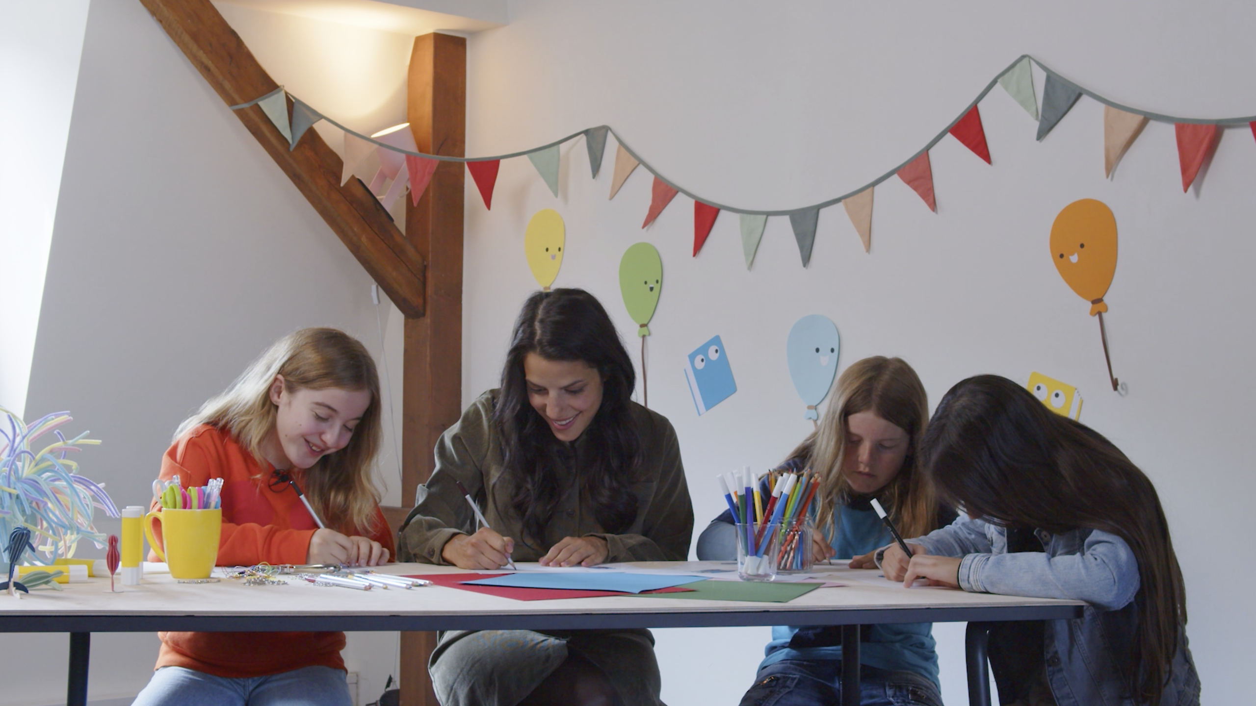 Das Bild zeigt eine Bastelrunde an einem Tisch. Auf dem Tisch liegen bunte Stifte, Papier und Bastelmaterial. Im Hintergrund hängen Wimpel und Luftballons mit Gesichtern als Dekoration an der Wand.