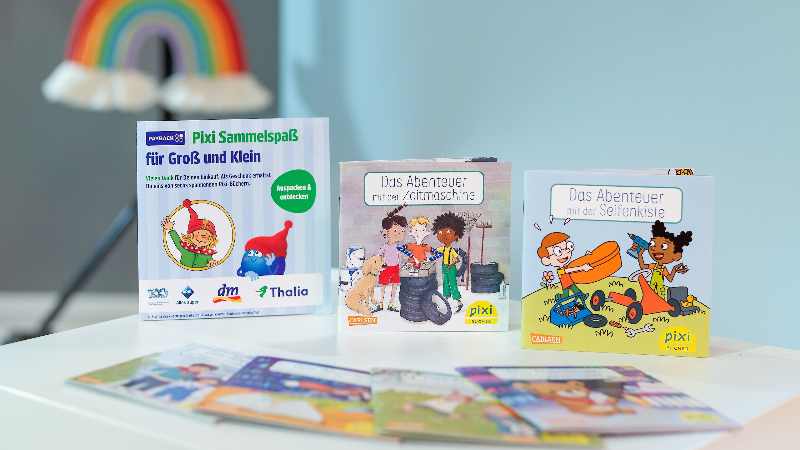 Das Bild zeigt drei Pixi-Bücher. Links steht ein Aufsteller mit „Pixi Sammelspaß für Groß und Klein“. In der Mitte ist das Buch „Das Abenteuer mit der Zeitmaschine“ mit Kindern, einem Hund und Reifen. Rechts steht „Das Abenteuer mit der Seifenkiste“ mit zwei Kindern, die eine Seifenkiste bauen. Im Hintergrund ist ein Regenbogen zu sehen.