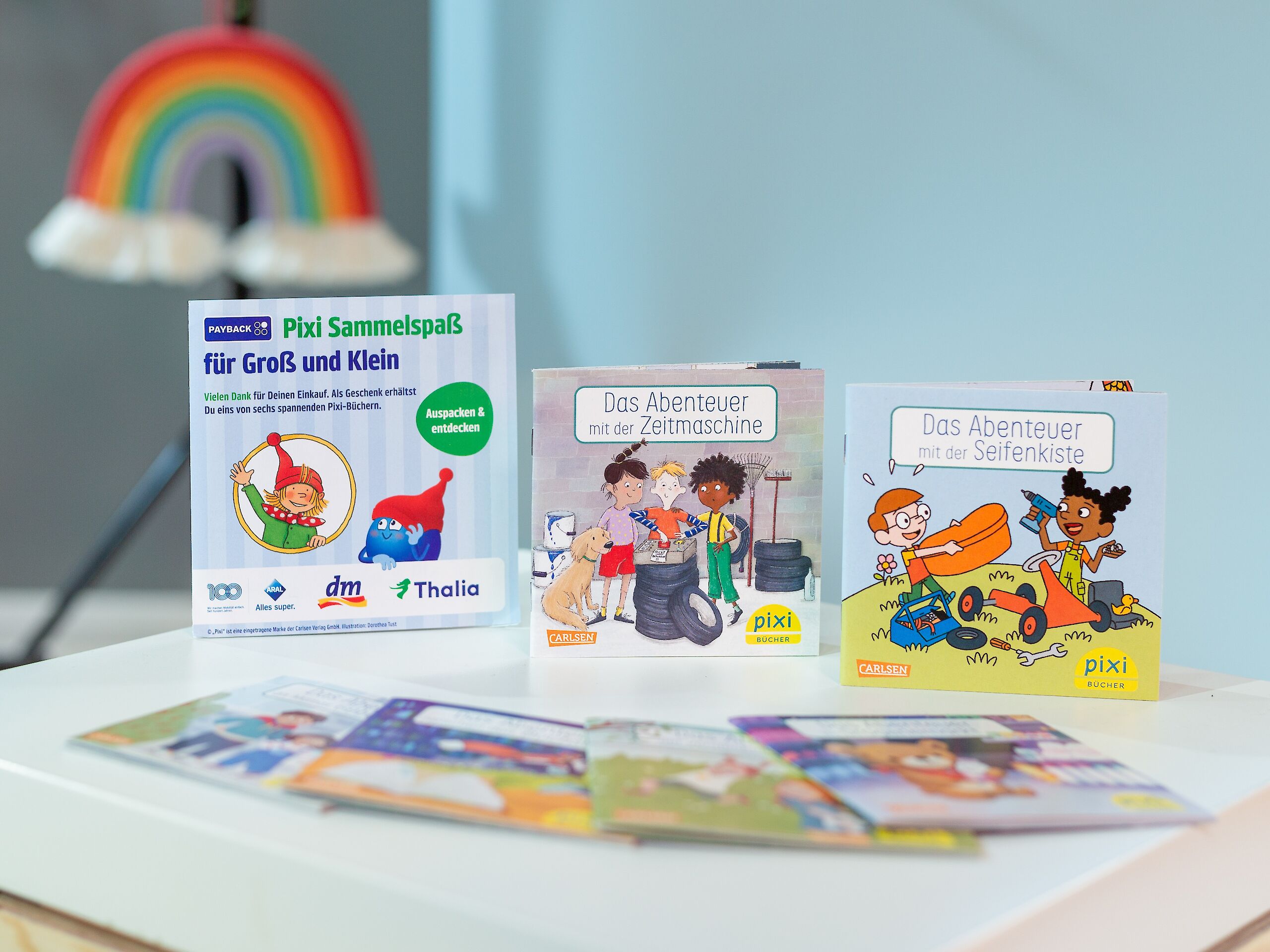 Das Bild zeigt drei Pixi-Bücher. Links steht ein Aufsteller mit „Pixi Sammelspaß für Groß und Klein“. In der Mitte ist das Buch „Das Abenteuer mit der Zeitmaschine“ mit Kindern, einem Hund und Reifen. Rechts steht „Das Abenteuer mit der Seifenkiste“ mit zwei Kindern, die eine Seifenkiste bauen. Im Hintergrund ist ein Regenbogen zu sehen.
