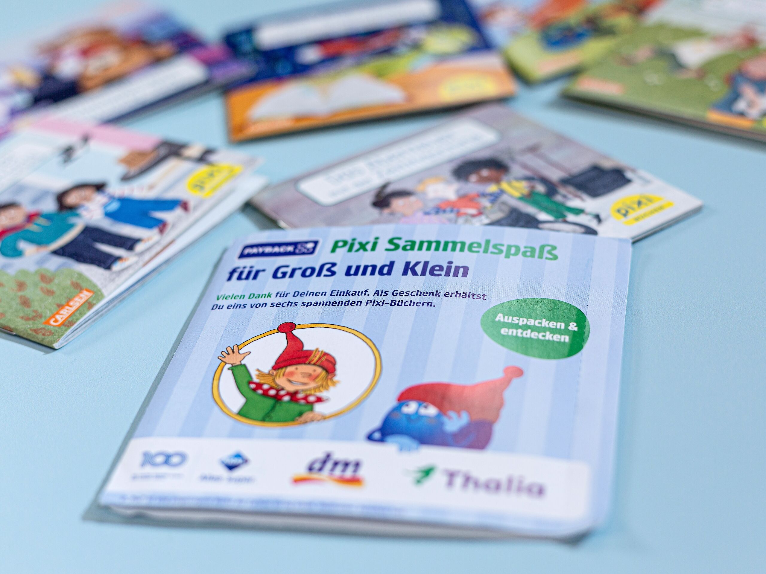 Das Bild zeigt mehrere Pixi-Bücher, die auf einem Tisch liegen. Im Vordergrund ist ein Aufsteller mit der Aufschrift „Pixi Sammelspaß für Groß und Klein“ zu sehen, daneben bunte Buchcover mit verschiedenen Motiven.