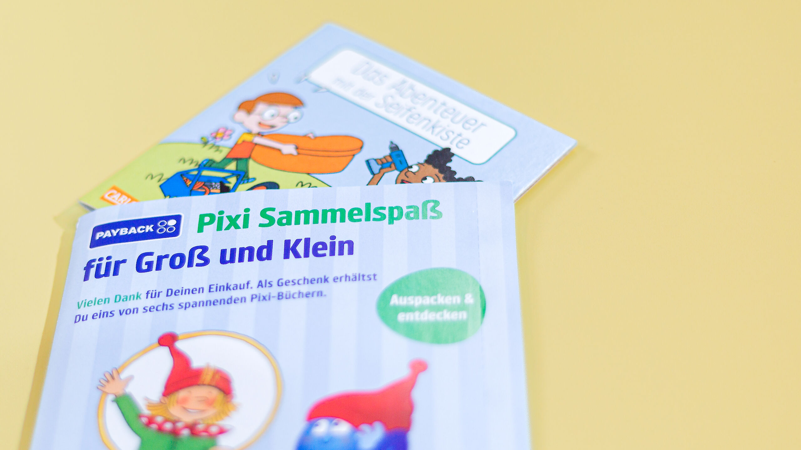 Das Bild zeigt die Überraschungsverpackung mit der Aufschrift „Pixi Sammelspaß für Groß und Klein“ und ein Pixi-Buch mit dem Titel „Das Abenteuer mit der Seifenkiste“. Beide liegen auf einem gelben Hintergrund.