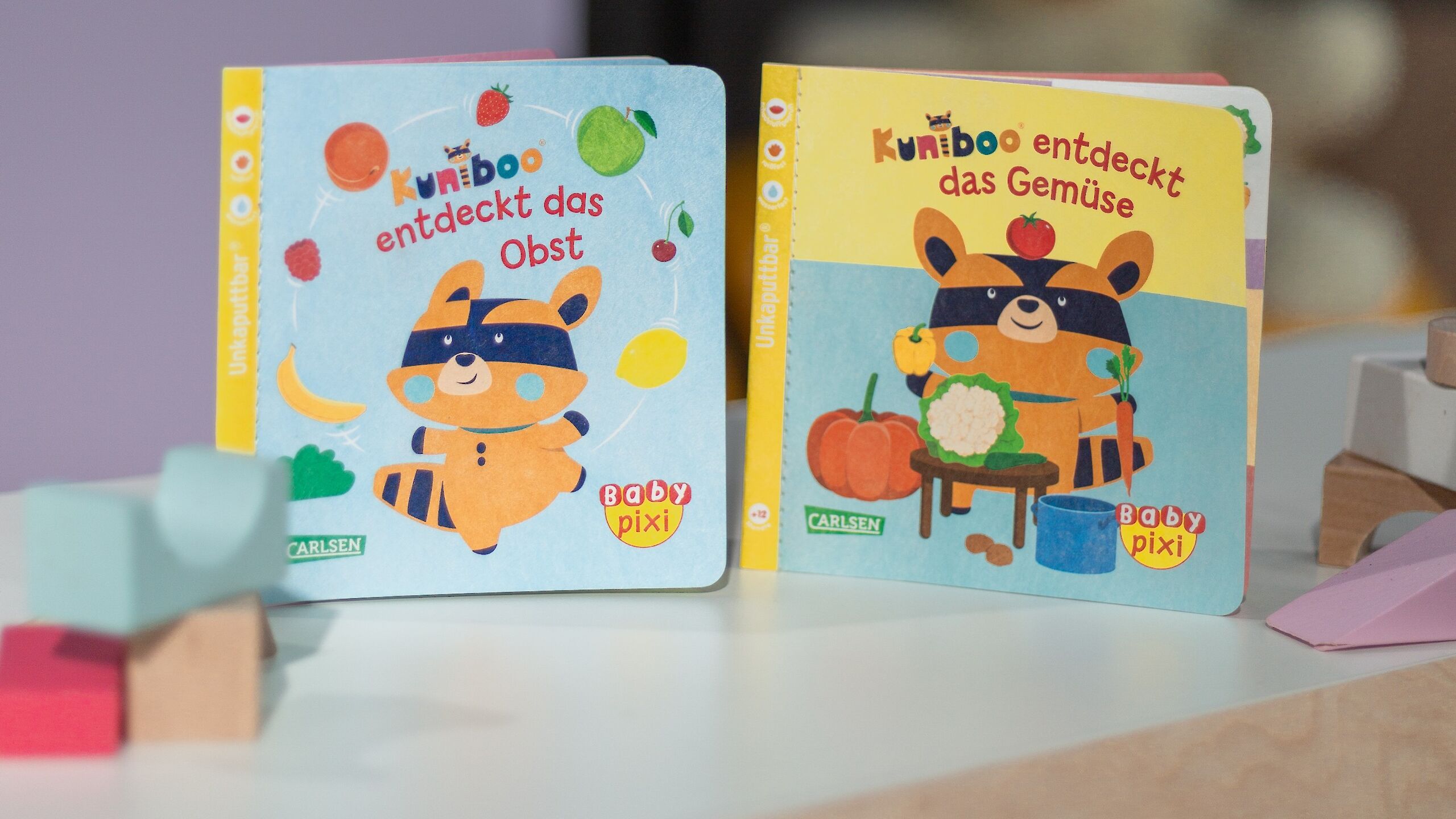 Das Bild zeigt zwei kleine Baby Pixi-Bücher aus der Reihe „Kuniboo“. Auf dem linken Buch steht „Kuniboo entdeckt das Obst“. Es zeigt einen Waschbären mit einer Augenmaske, der von verschiedenen Obstsorten wie einer Banane, einem Apfel und einer Erdbeere umgeben ist. Das Cover ist hellblau.  Das rechte Buch heißt „Kuniboo entdeckt das Gemüse“. Auch hier ist der Waschbär zu sehen, diesmal mit Gemüse wie einem Kürbis, Blumenkohl und einer Tomate. Das Cover ist gelb.  Im Hintergrund sieht man eine verschwommene Szene mit Wimpeln und einem Teddy. Die Bücher stehen auf einem Tisch, auf dem auch bunte Holzklötze liegen.