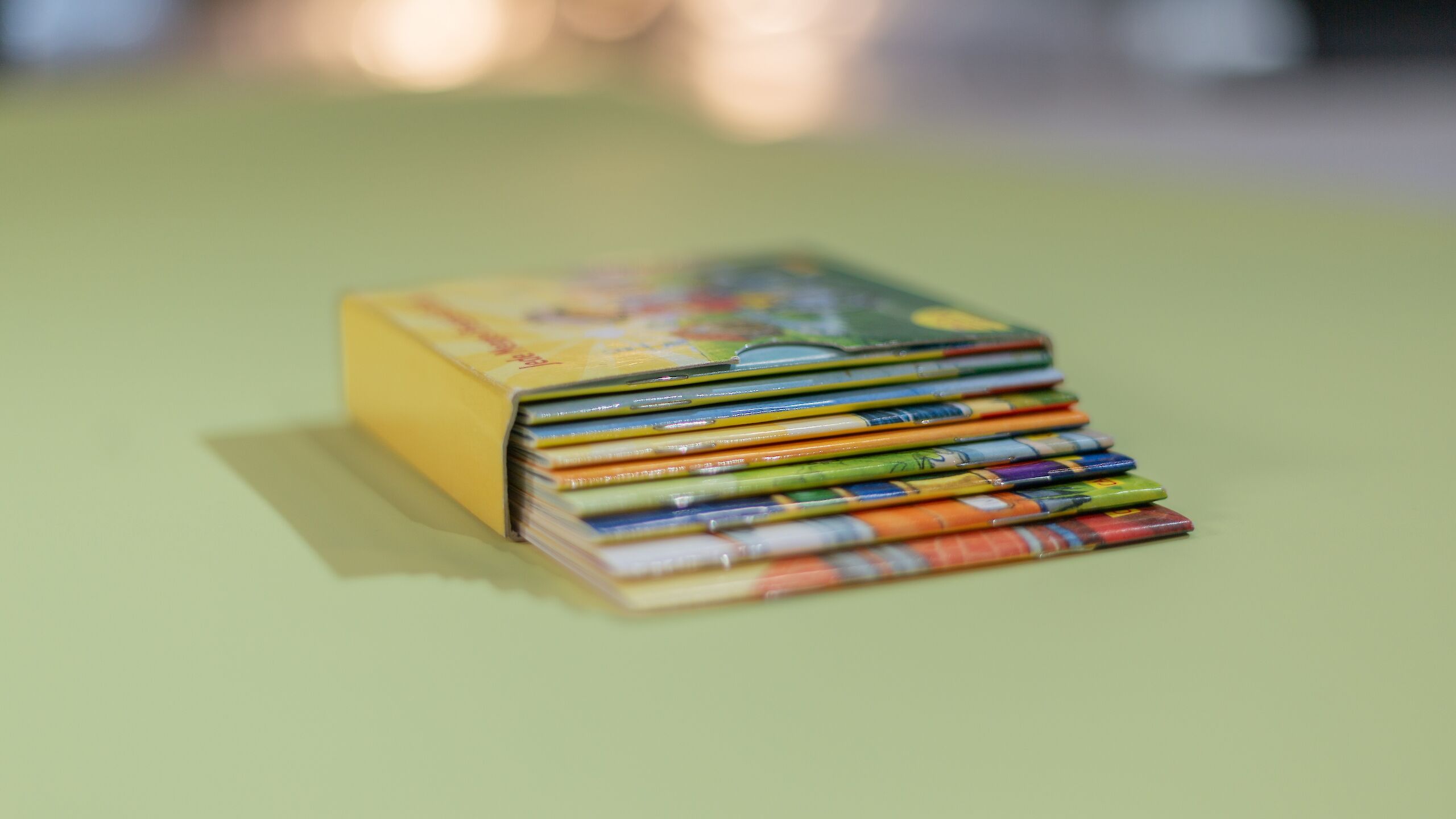Das Bild zeigt einen Stapel bunter Pixi-Bücher in einer gelben Box. Die Bücher liegen auf einem grünen Untergrund.