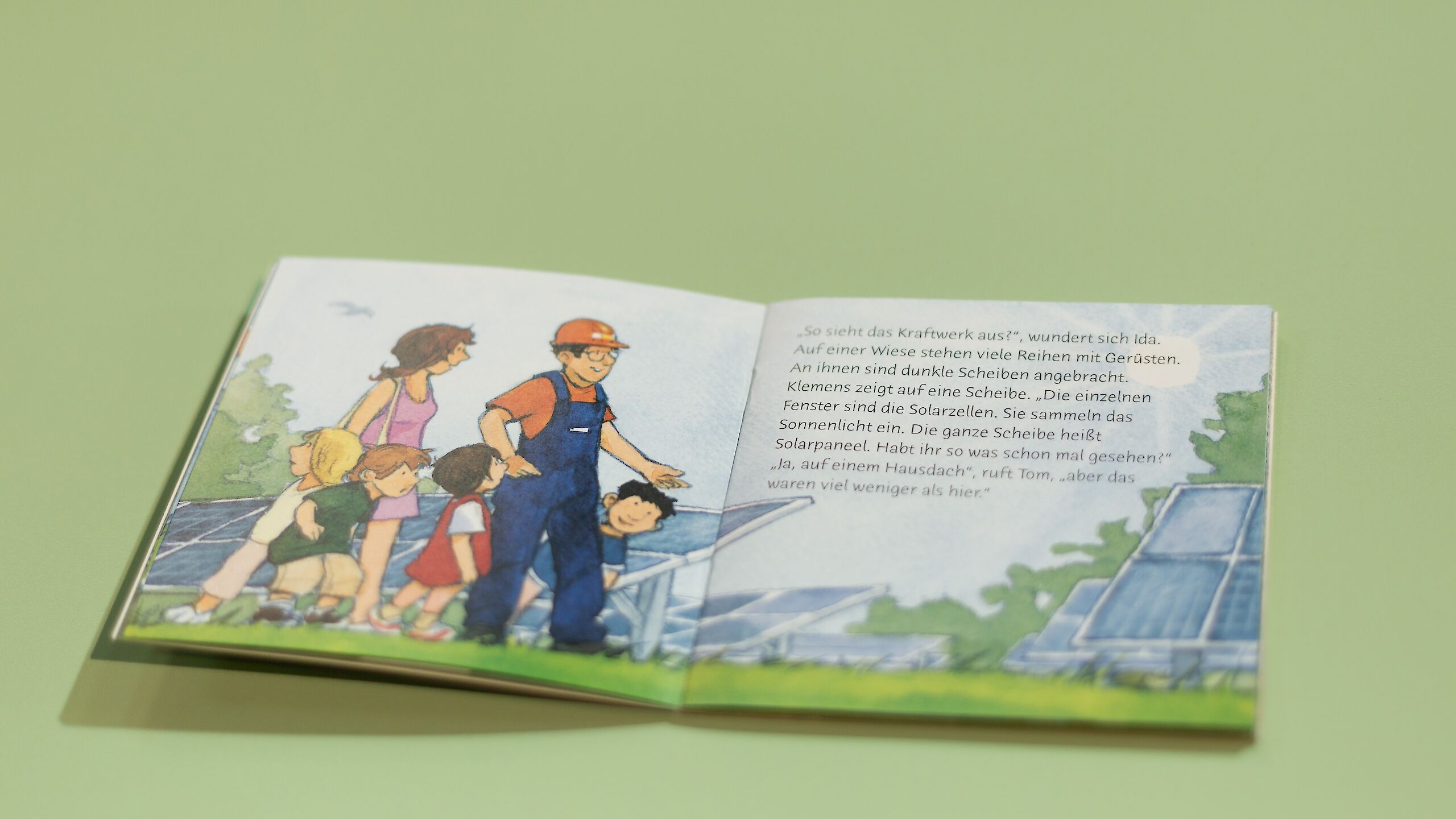Ein aufgeschlagenes Pixi-Buch liegt auf einem grünen Tisch. Auf der linken Seite stehen Kinder und zwei Erwachsene vor Solarzellen.