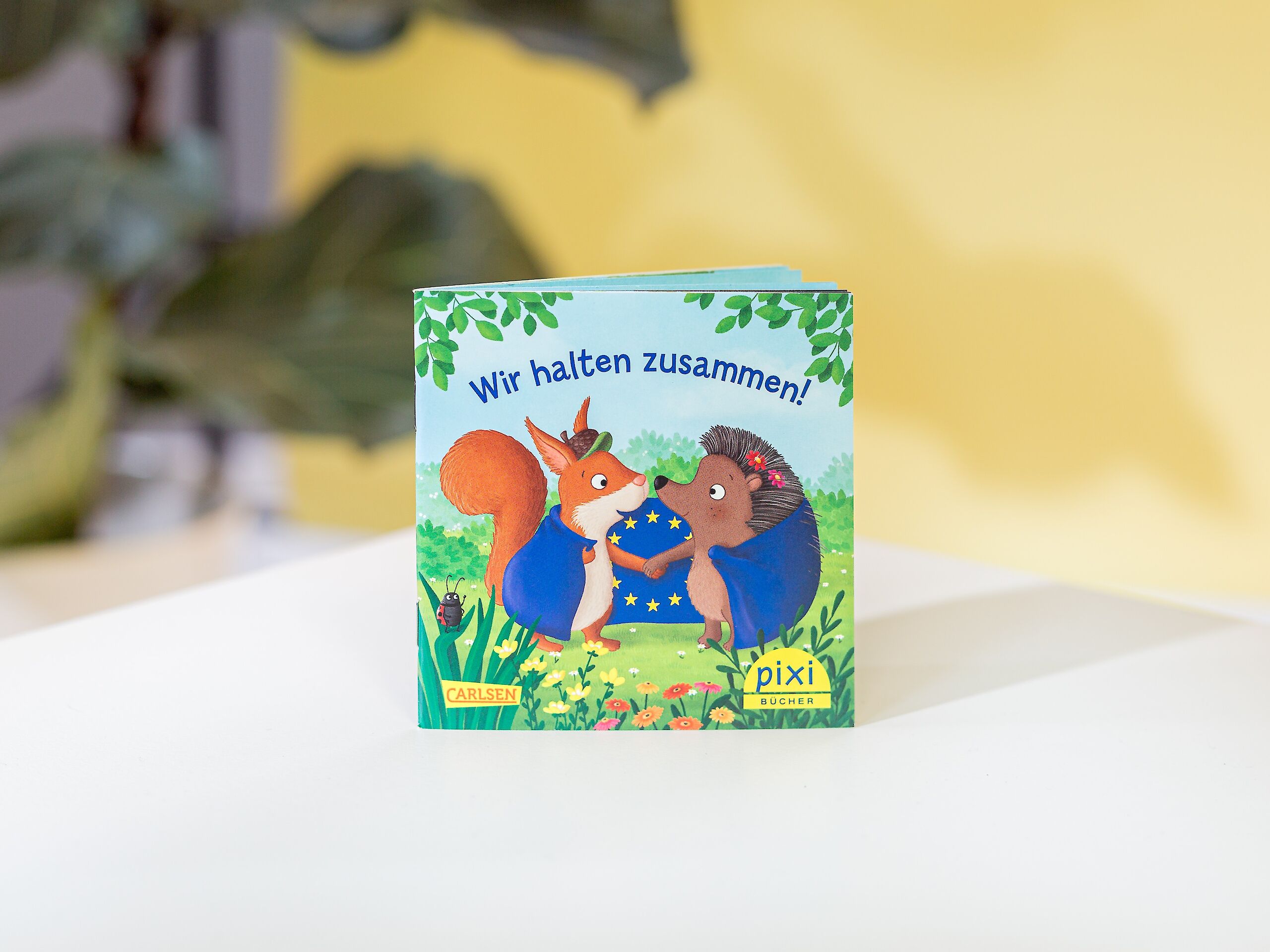 Auf dem Bild sieht man ein Pixi-Buch mit dem Titel „Wir halten zusammen!“. Auf dem Titelbild sind ein Eichhörnchen und ein Igel, die sich an den Händen halten. Der Hintergrund zeigt eine Wiese mit Blumen.
