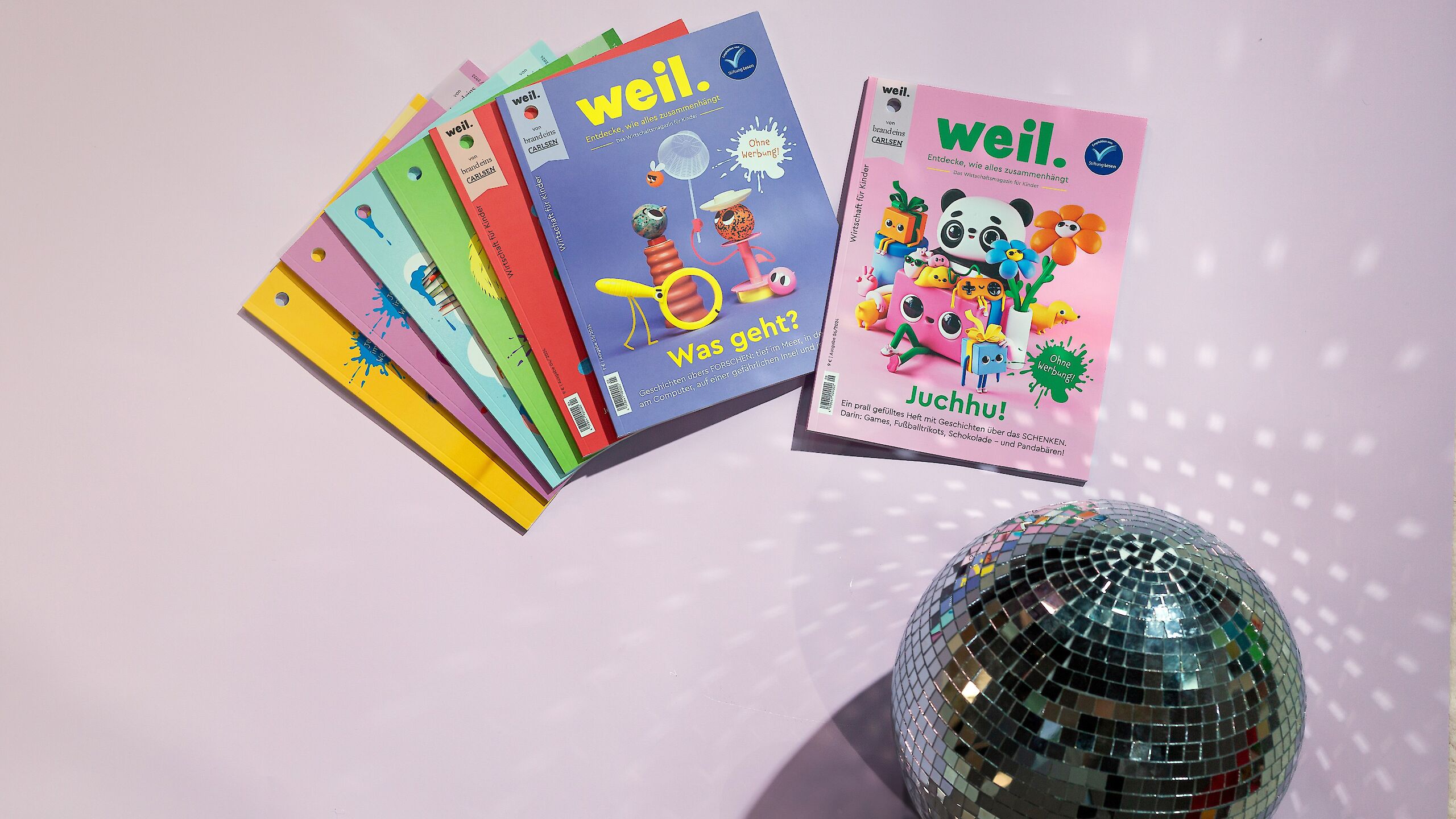Das Bild zeigt mehrere bunte Ausgaben des Magazins „weil.“. Zwei Hefte liegen vorne: Eines mit dem Titel „Was geht?“ und eines mit „Juchhu!“. Im Vordergrund ist eine silberne Discokugel zu sehen.