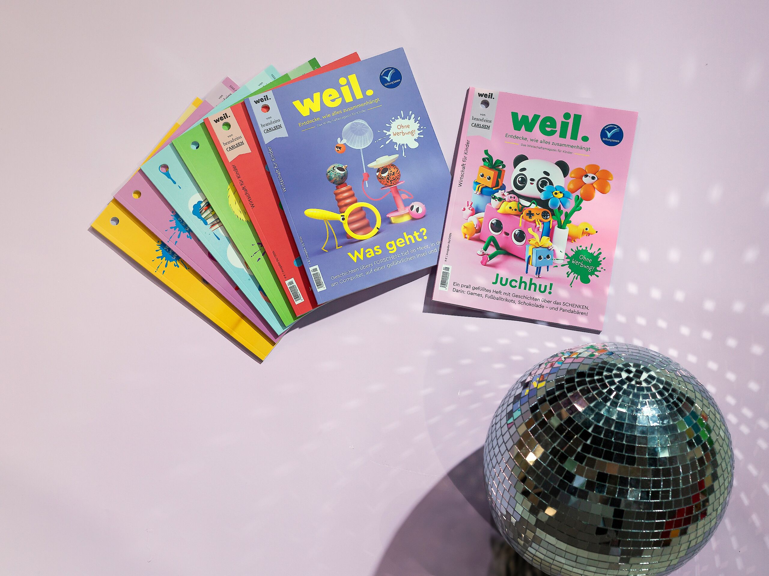 Das Bild zeigt mehrere bunte Ausgaben des Magazins „weil.“. Zwei Hefte liegen vorne: Eines mit dem Titel „Was geht?“ und eines mit „Juchhu!“. Im Vordergrund ist eine silberne Discokugel zu sehen.