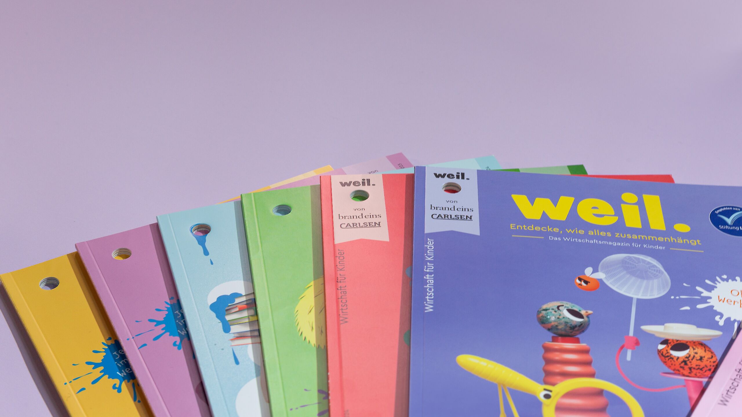 Das Bild zeigt mehrere bunte Ausgaben des Magazins „weil.“. Im Vordergrund ist ein Heft mit lila Cover. Die Hefte sind fächerförmig angeordnet.