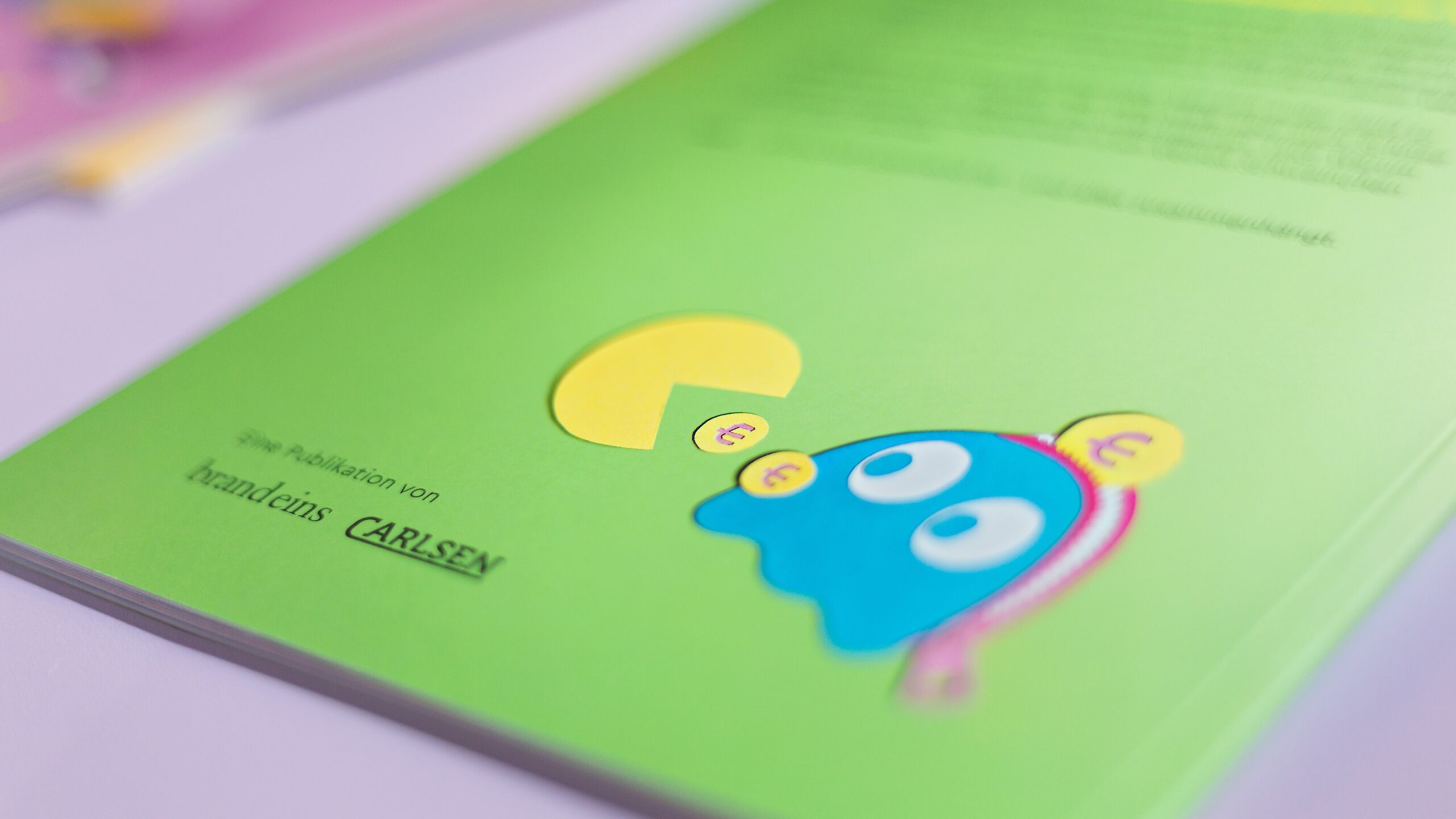 Das Bild zeigt eine grüne Seite mit einer Illustration: Ein gelber Kreis ähnelt Pac-Man und frisst Münzen mit Euro-Zeichen. Rechts daneben ist ein blauer Charakter mit großen Augen. Unten steht „Eine Publikation von brand eins und CARLSEN“.