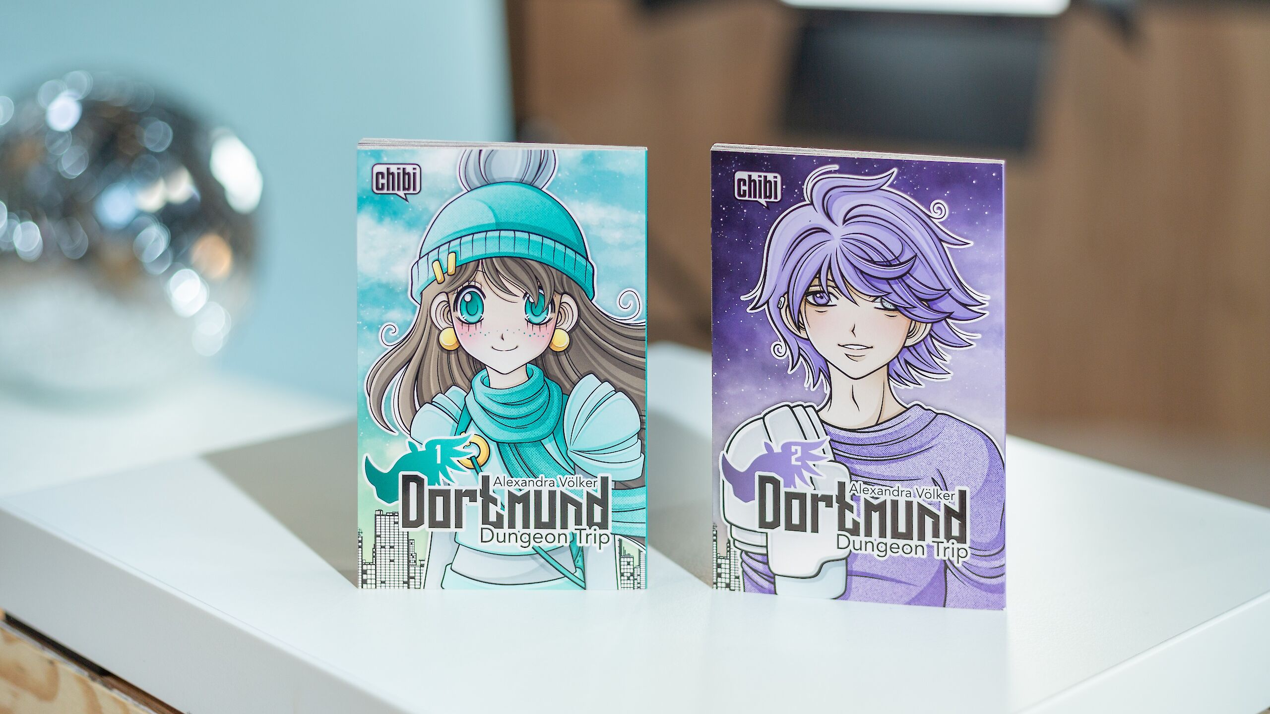 Auf dem Bild sind zwei Manga-Bücher mit dem Titel „Dortmund Dungeon Trip“ zu sehen. Das linke Cover zeigt ein Mädchen mit Mütze und Schal vor einem blauen Hintergrund, das rechte einen Charakter mit lila Haaren vor einem violetten Sternenhimmel.