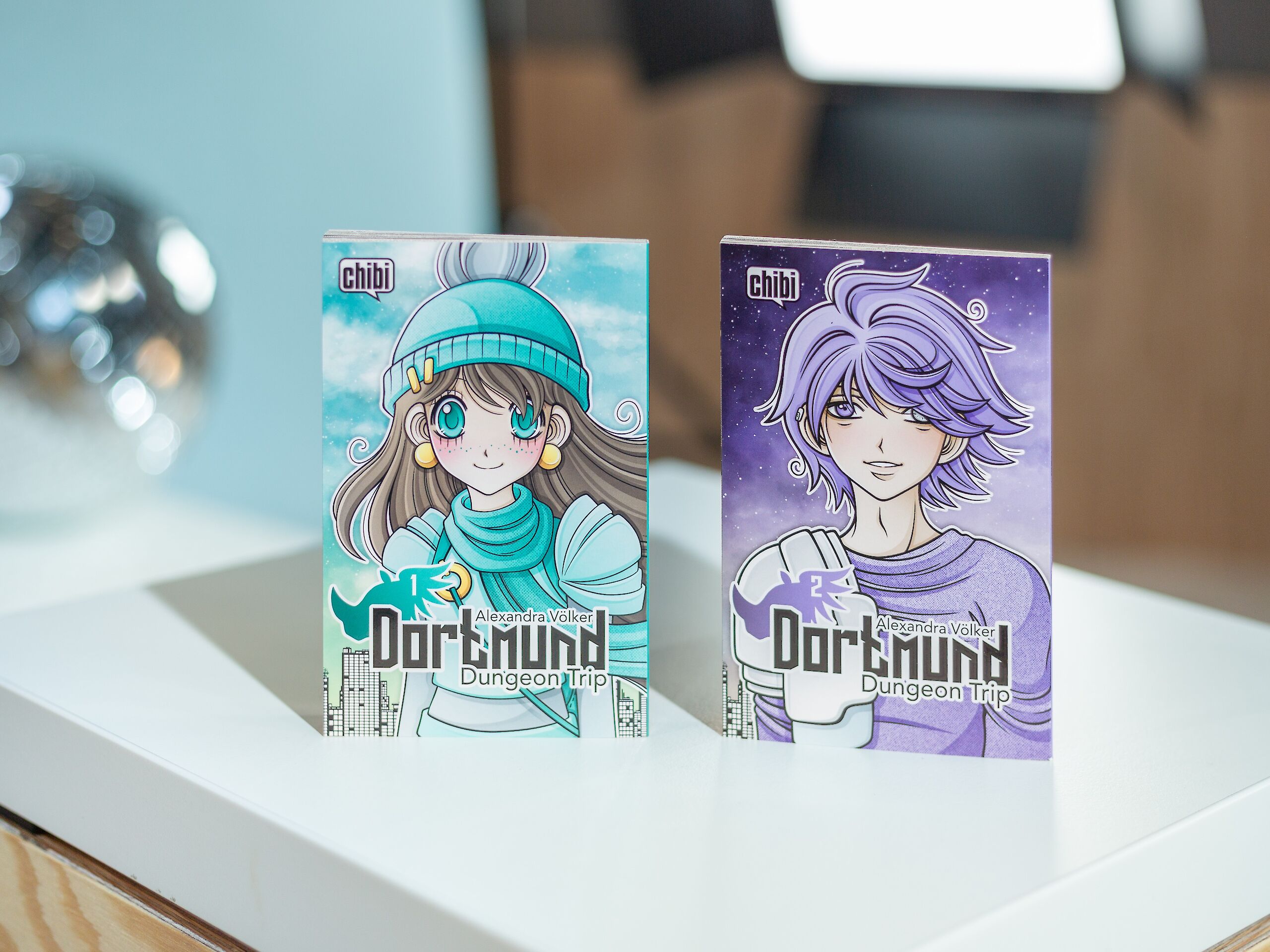 Auf dem Bild sind zwei Manga-Bücher mit dem Titel „Dortmund Dungeon Trip“ zu sehen. Das linke Cover zeigt ein Mädchen mit Mütze und Schal vor einem blauen Hintergrund, das rechte einen Charakter mit lila Haaren vor einem violetten Sternenhimmel.