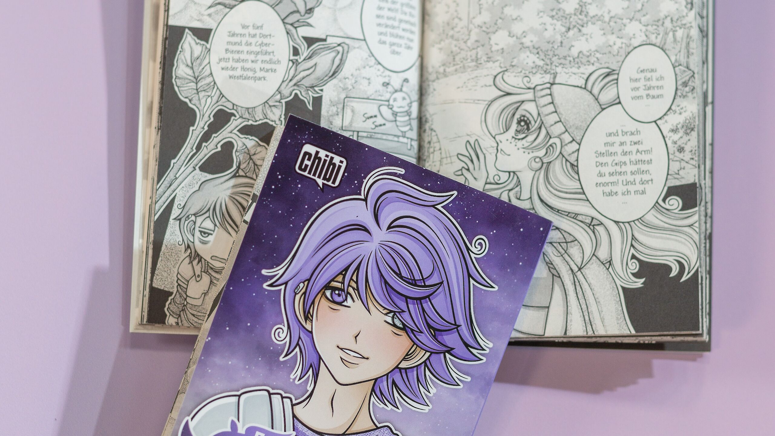 Auf dem Bild liegt ein aufgeschlagenes Manga-Buch mit schwarz-weißen Zeichnungen vor einem lila Hintergrund. Davor liegt das Cover eines Manga-Bandes mit dem Titel „Dortmund Dungeon Trip“.