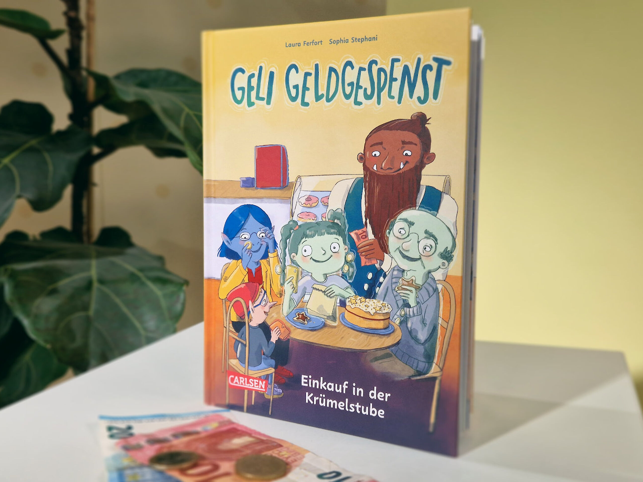 Das Buch "Geli Geldgespenst – Einkauf in der Krümelstube" auf einem weißen Tisch. Daneben liegen Euroscheine und -münzen.